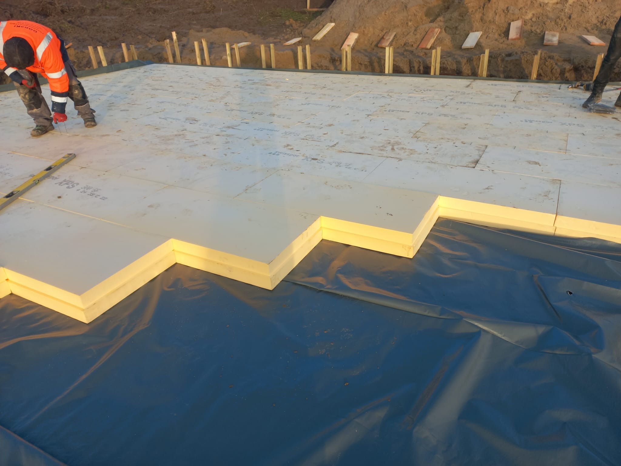 Układanie izolacji XPS na folii hydroizolacyjnej pod fundament. Widoczny pracownik w odblaskowej kurtce i poziomica. Drewniane szalunki w tle.