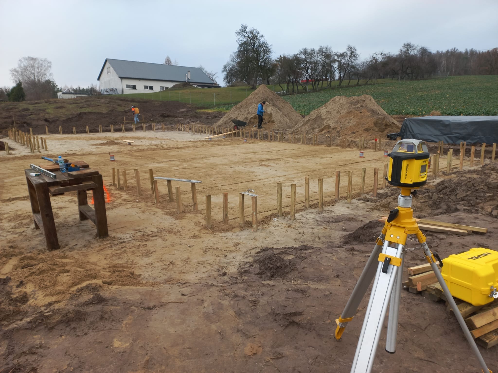 Wytyczanie fundamentów pod budynek z użyciem niwelatora laserowego. W tle widoczny dom i pracownicy z taczką. Drewniane słupki wyznaczają obrys.