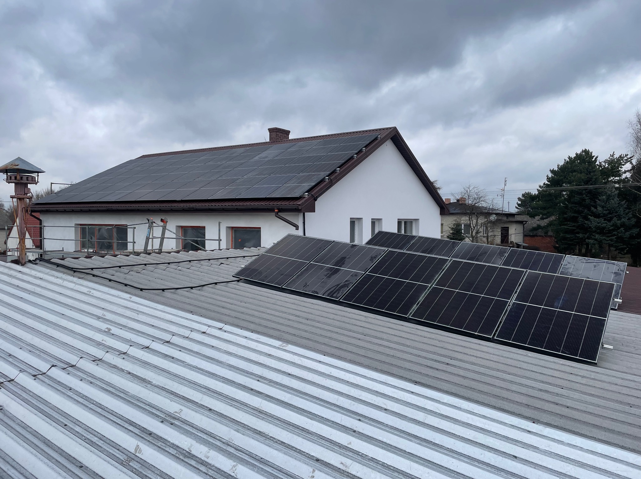 Panele fotowoltaiczne na dachu domu jednorodzinnego, widok z dachu sąsiedniego budynku. Szare niebo w tle. Widoczna drabina i komin.