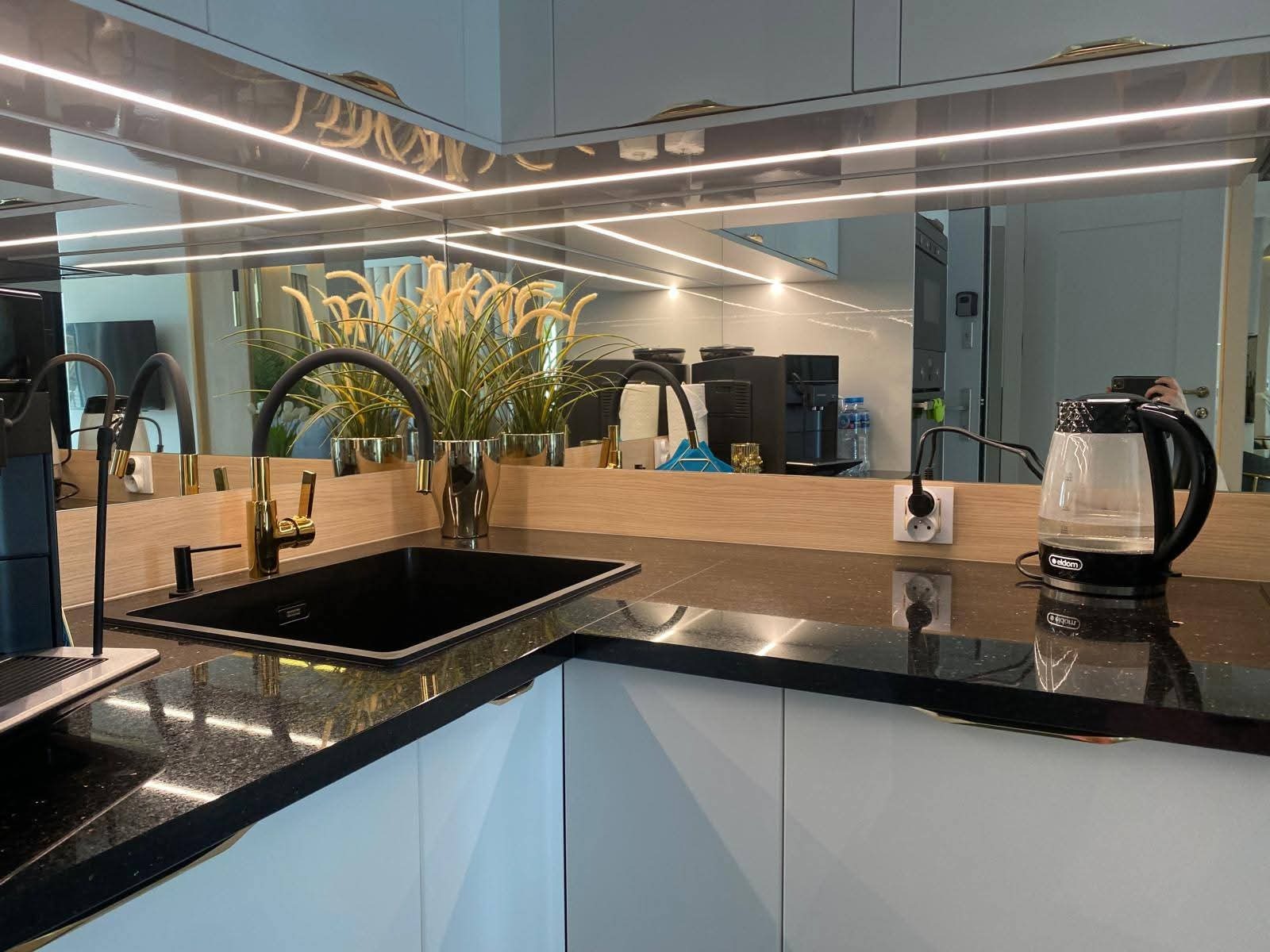 Czarny, błyszczący blat kamienny z wbudowanym zlewem i złotą baterią w nowoczesnej kuchni z lustrzanym panelem nad blatem i oświetleniem LED.