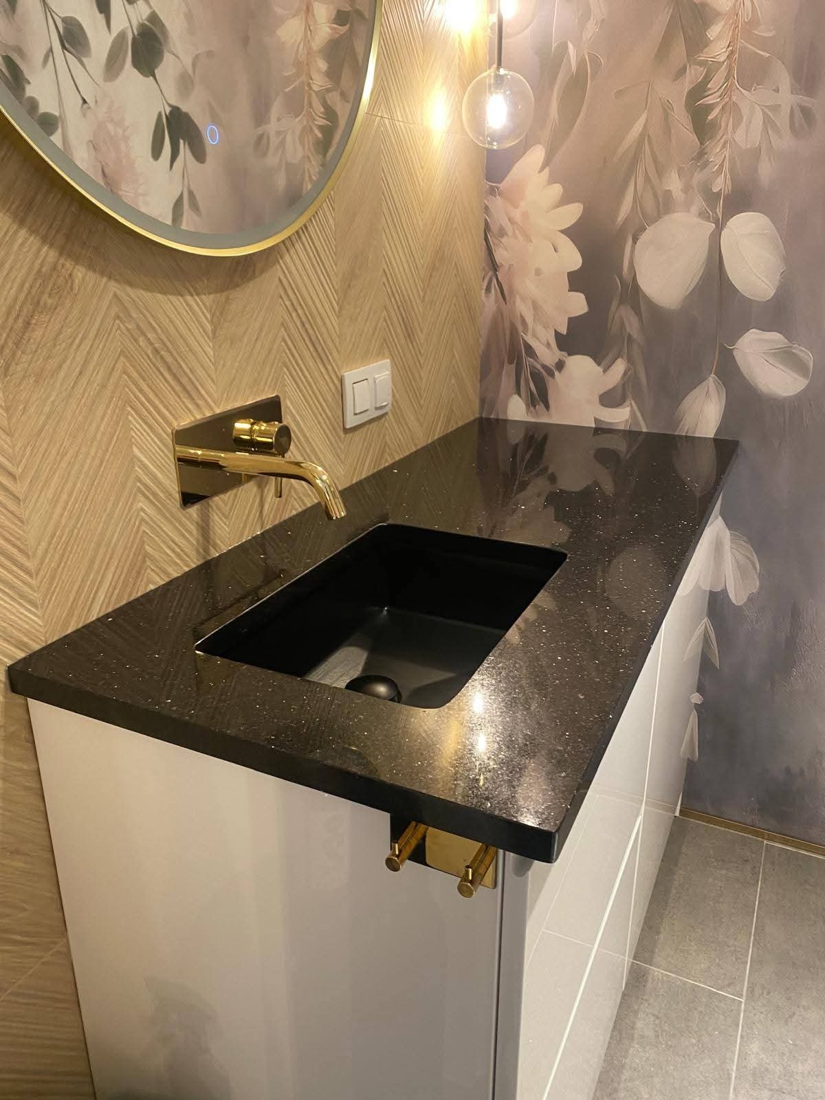 Błyszczący, czarny blat kamienny z brokatem, umywalka wpuszczana, złota bateria i uchwyty, na tle wzorzystej ściany i okrągłego lustra z podświetleniem LED.