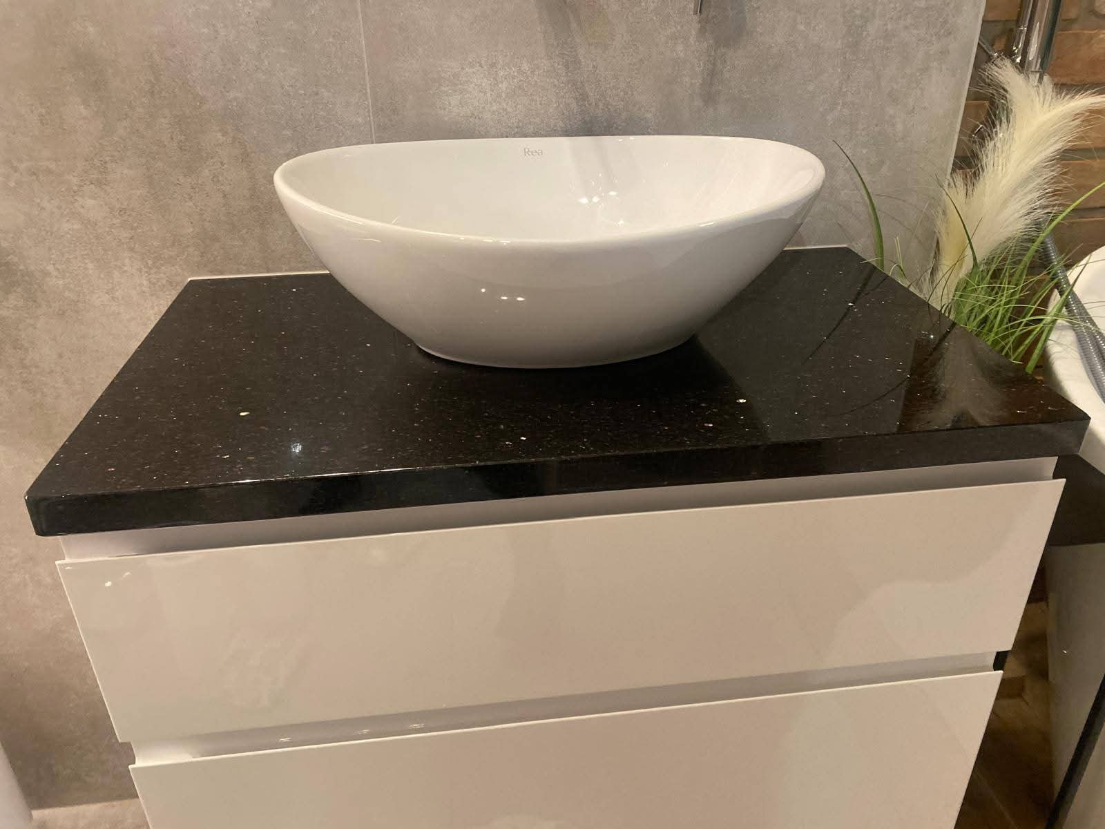 Czarny blat kamienny z umywalką Rea na białej szafce łazienkowej. Umywalka owalna, biała. W tle szara ściana i dekoracyjne trawy.