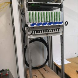 M_Pro_Sys - Szafa telekomunikacyjna z panelami światłowodowymi i okablowaniem, widok z boku, na tle ściany i biurka. Elementy w kolorach zielonym, niebieskim i czarnym.