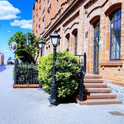 ENGLISH NAVIGATOR SPÓŁKA Z OGRANICZONĄ ODPOWIEDZIALNOŚCIĄ - Ceglany budynek z oknami łukowymi i czarnymi lampami ulicznymi, otoczony zielenią. Wejście z kilkoma schodami. Błękitne niebo w tle.