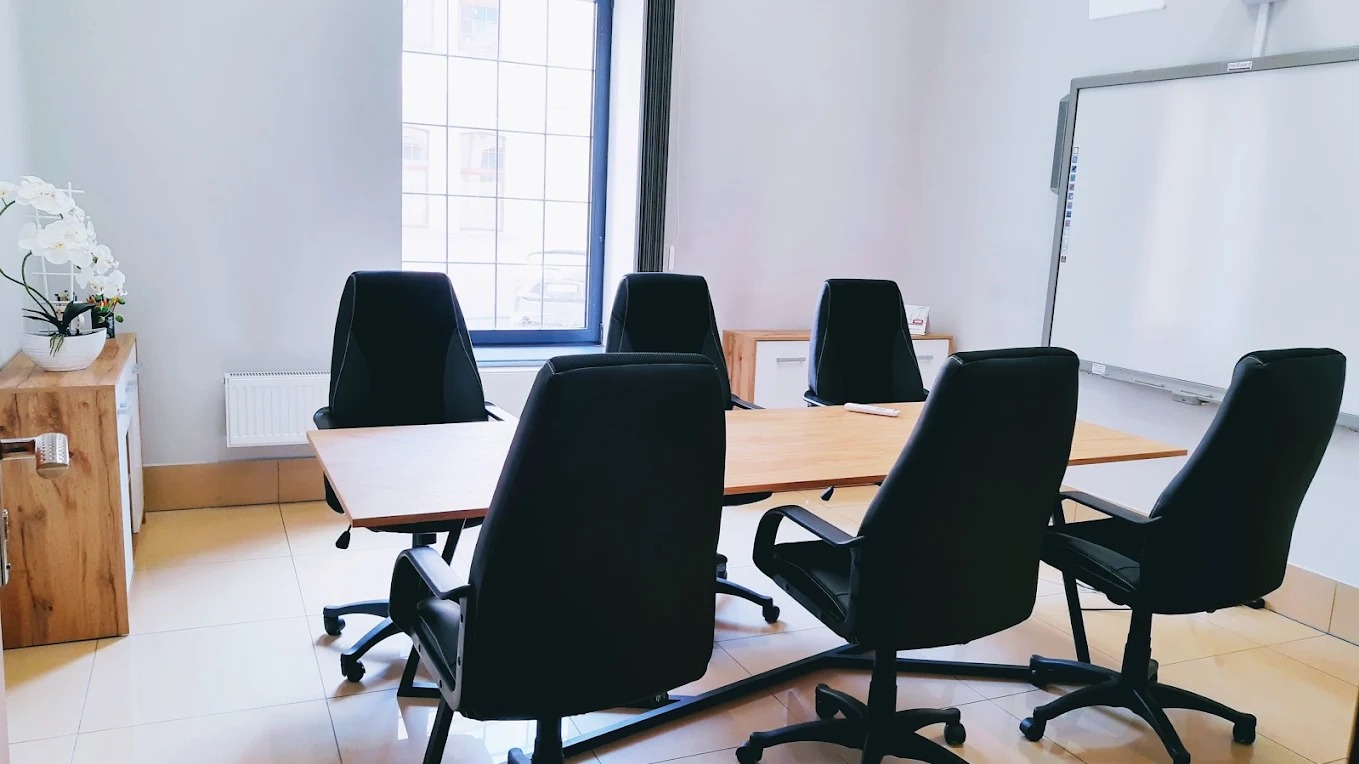 Sala lekcyjna z dużym oknem, stołem konferencyjnym i krzesłami obrotowymi. Na ścianie biała tablica suchościeralna. W rogu szafka z kwiatami.