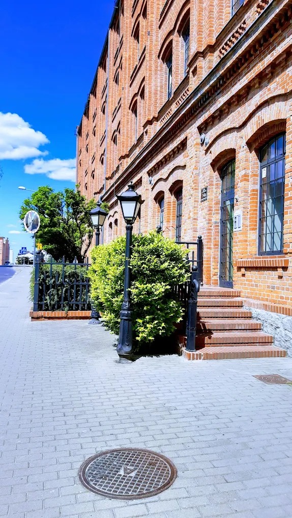 Ceglany budynek z oknami łukowymi i czarnymi lampami ulicznymi, otoczony zielenią. Wejście z kilkoma schodami. Błękitne niebo w tle.