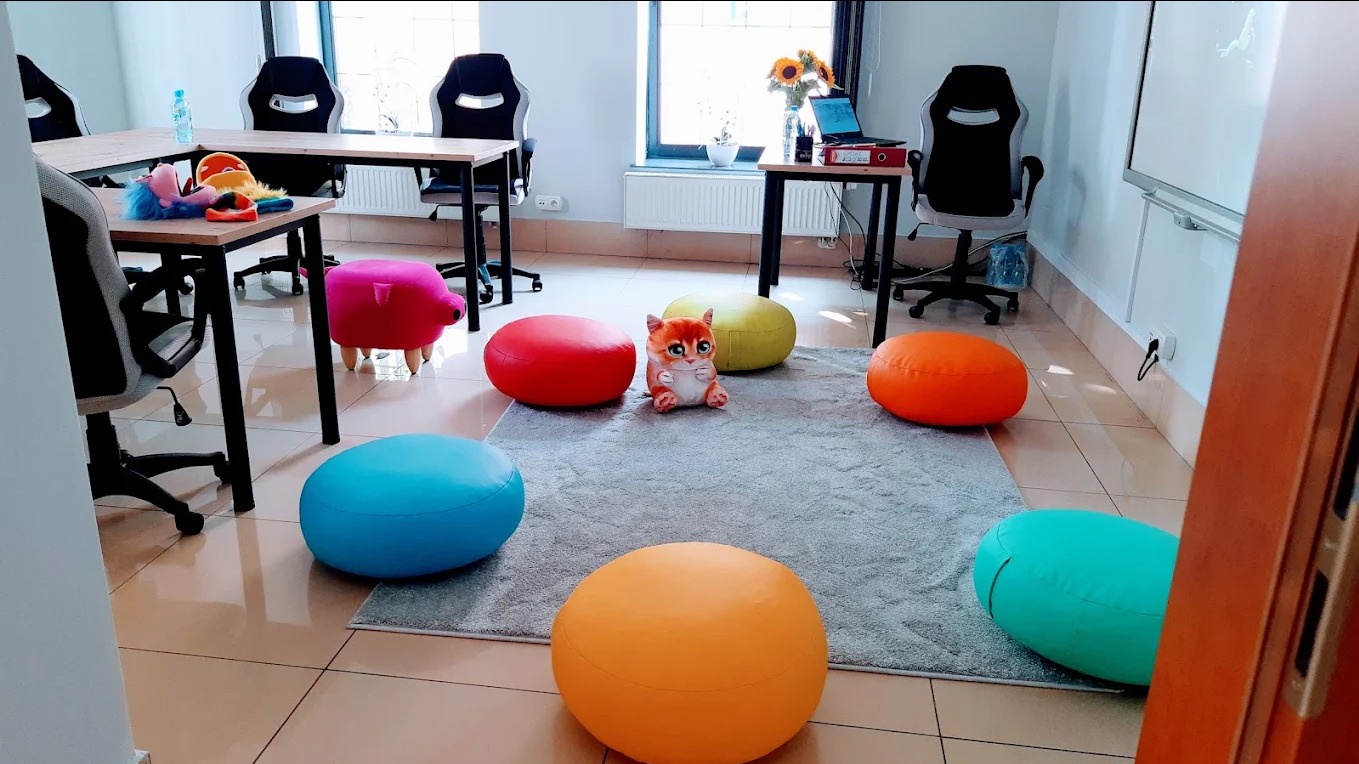 Kolorowe pufy i zabawki w jasnej sali lekcyjnej z biurkami i krzesłami. Przy oknie laptop i słoneczniki. Przyjazna przestrzeń edukacyjna dla dzieci.