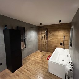 New Invest - Nowoczesna łazienka z drewnopodobną podłogą, szarą ścianą i kabiną prysznicową typu walk-in. Biała wanna z czarną armaturą i minimalistyczne oświetlenie.