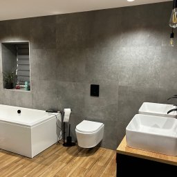 New Invest - Nowoczesna łazienka z wanną, toaletą podwieszaną i dwiema umywalkami nablatowymi. Szare płytki imitujące kamień, drewnopodobna podłoga, czarne akcenty.