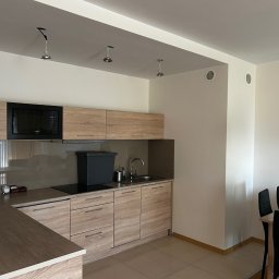New Invest - Jasna kuchnia z drewnianymi szafkami, czarną mikrofalówką i trzema punktami oświetleniowymi na suficie. Widoczny fragment stołu z krzesłami.