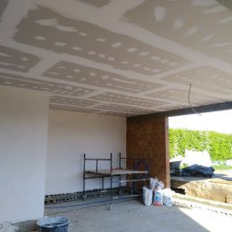 KRZYSZTOF KUSTRA Usługi Ogólno-Budowlane - Wnętrze w trakcie prac wykończeniowych: sufit z płyt karton-gips, częściowo wyszpachlowane ściany, rusztowanie i worki z zaprawą. Widok na zewnątrz przez otwartą przestrzeń.