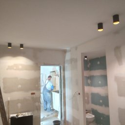 KRZYSZTOF KUSTRA Usługi Ogólno-Budowlane - Wnętrze w trakcie prac wykończeniowych: ściany przygotowane pod malowanie, widoczne punkty oświetleniowe, drabina i pracownik w tle. Wnętrze w stanie surowym.