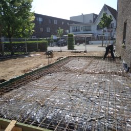 KRZYSZTOF KUSTRA Usługi Ogólno-Budowlane - Zbrojenie fundamentów pod budowę domu jednorodzinnego. Siatka zbrojeniowa na folii, drewniane szalunki, widoczny fragment budynku w tle. Prace przygotowawcze.