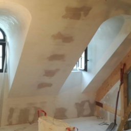 KRZYSZTOF KUSTRA Usługi Ogólno-Budowlane - Skośne poddasze ze świeżo nałożonymi gładziami, widoczne łaty na suficie i ścianach. Okna łukowe w czarnych ramach. Przy ścianie narzędzia i torba budowlana.