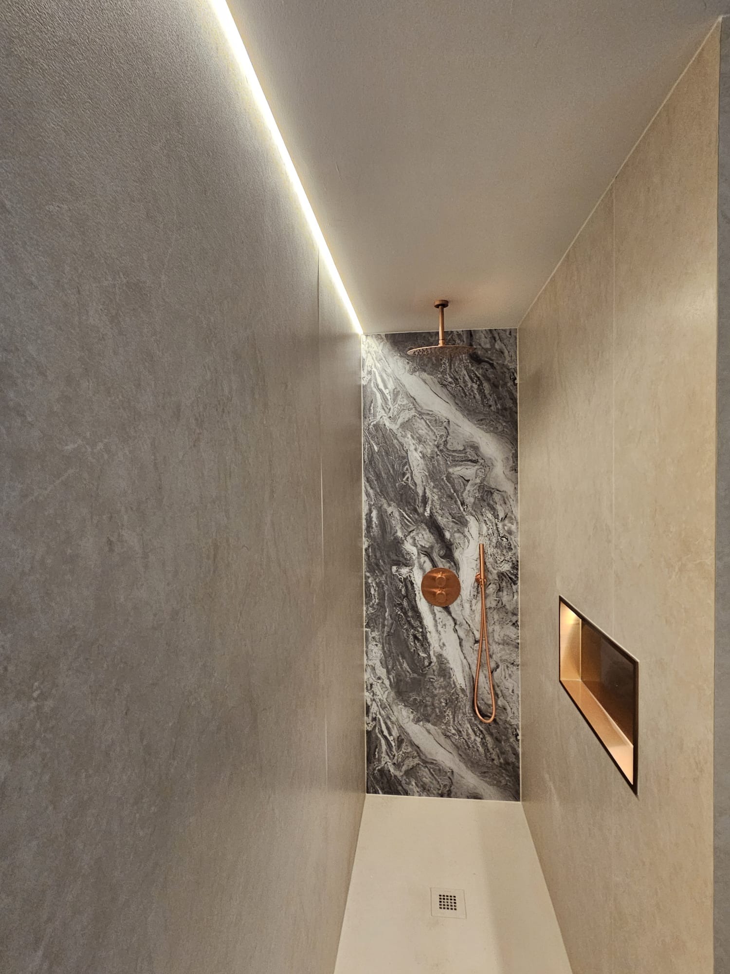 Nowoczesna kabina prysznicowa z marmurową ścianą akcentową, miedzianą armaturą i subtelnym oświetleniem LED. Minimalistyczny design z wnęką ścienną.