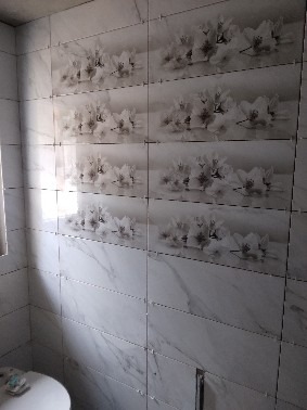 Białe, błyszczące płytki z motywem kwiatowym na ścianie łazienki, ułożone w rzędach. Widoczny fragment toalety i narożnik pomieszczenia. Delikatne światło.