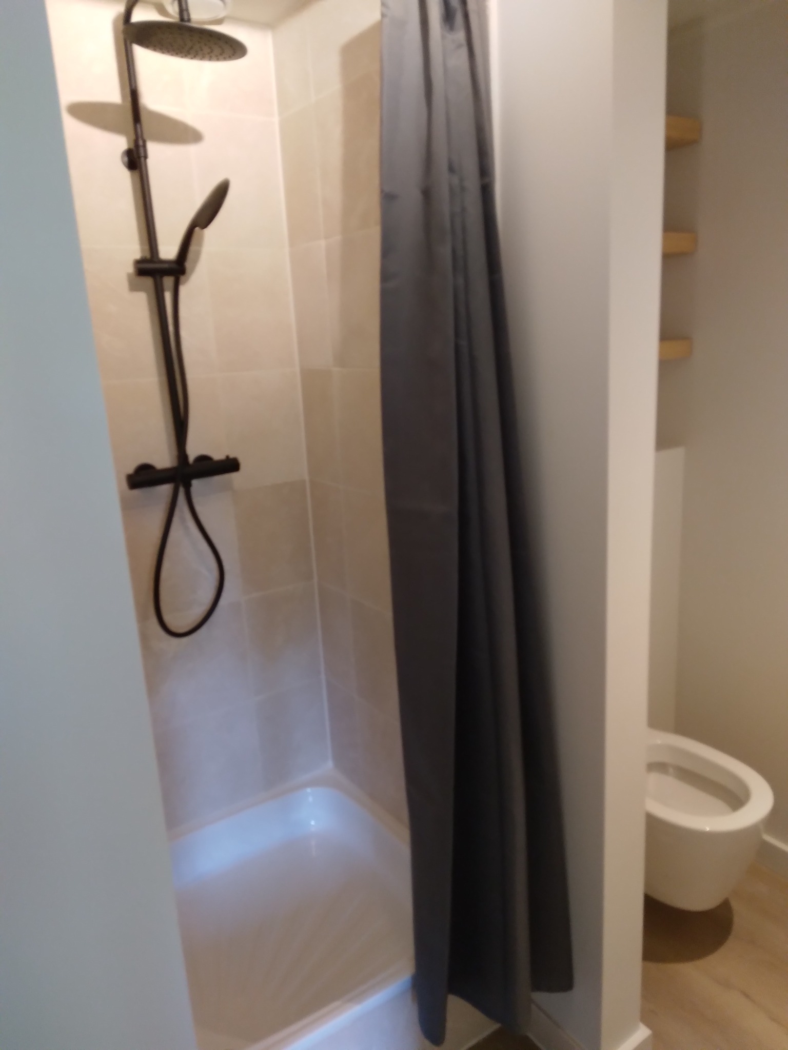 Nowoczesna kabina prysznicowa z deszczownicą i szarą zasłoną, widok na toaletę i półki w tle. Jasne płytki i minimalistyczny design.