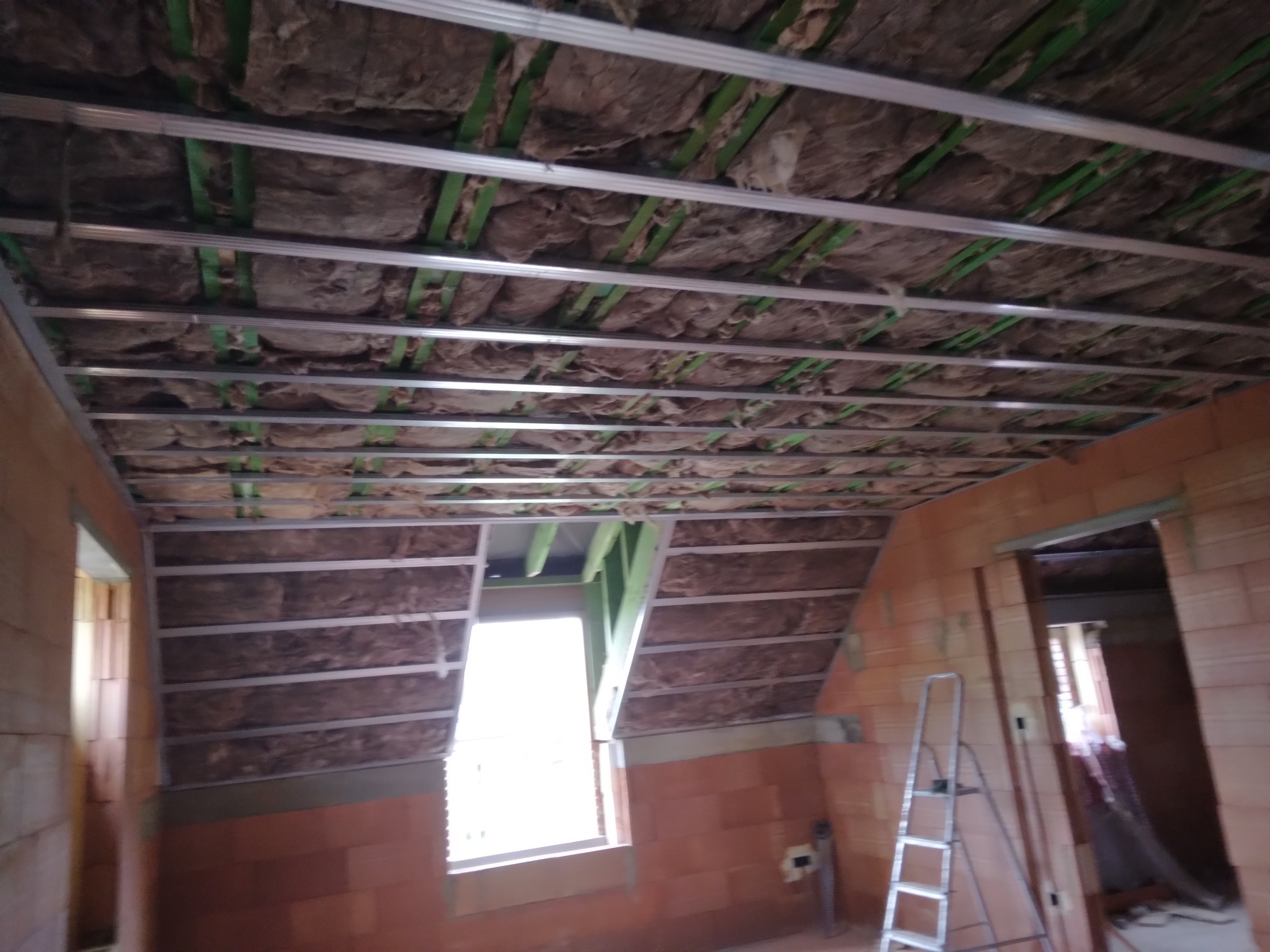 Poddasze z izolacją z wełny mineralnej, widoczne metalowe profile sufitowe i fragment ściany z cegły. W tle okno i drabina.