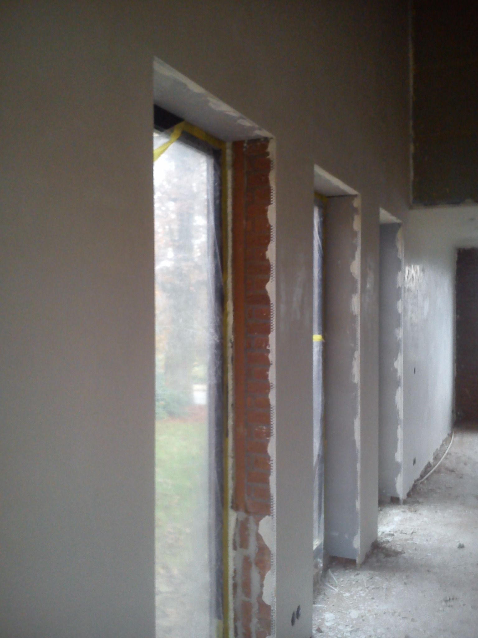 Ściana po szpachlowaniu przy oknie z widoczną cegłą i taśmą malarską. Widoczne ślady po gładzi. Prace wykończeniowe w surowym wnętrzu.