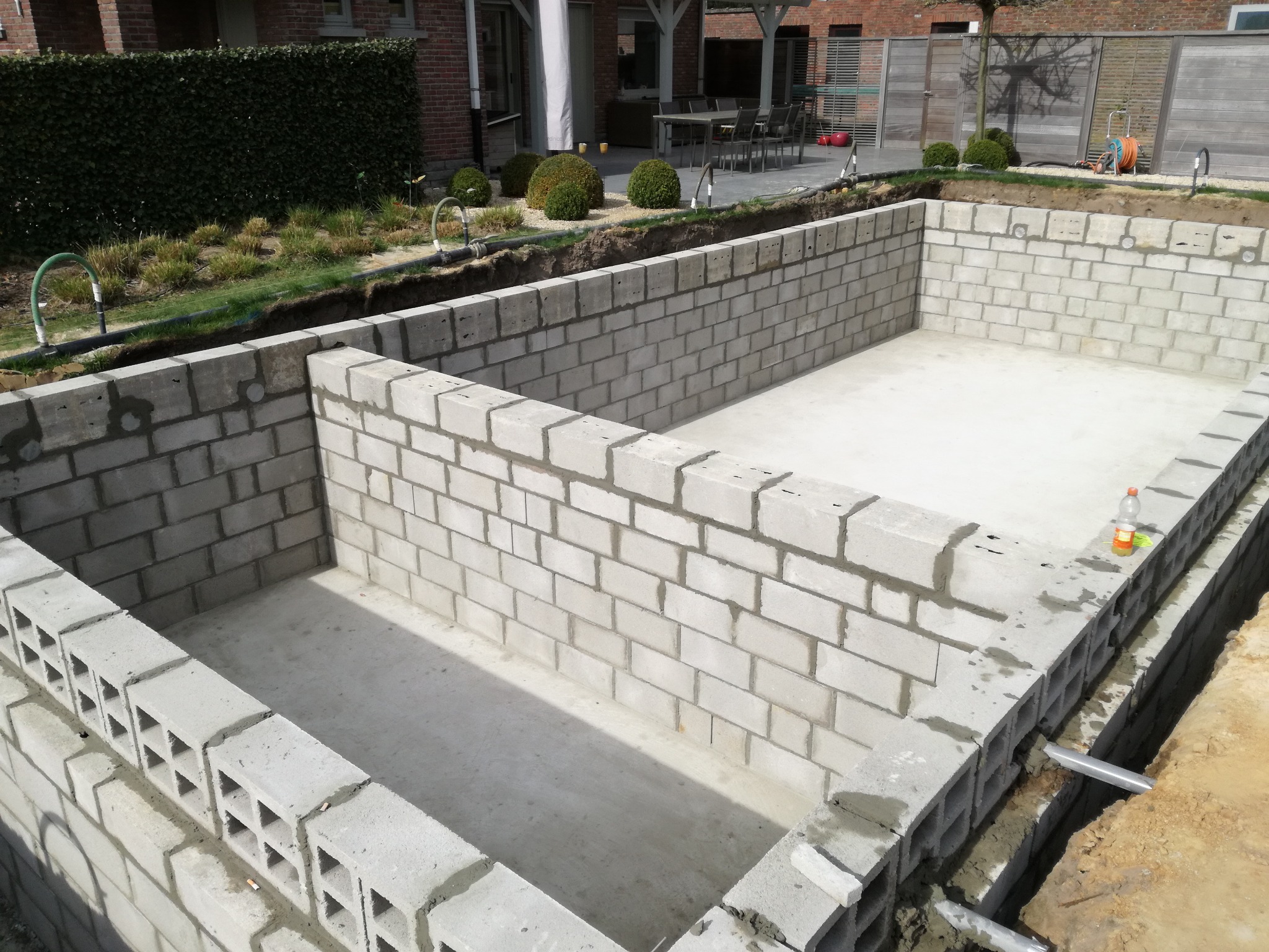 Murowana konstrukcja z betonowych bloczków na zewnątrz, prawdopodobnie fundament pod basen. Widoczne elementy instalacji i otoczenie ogrodu.