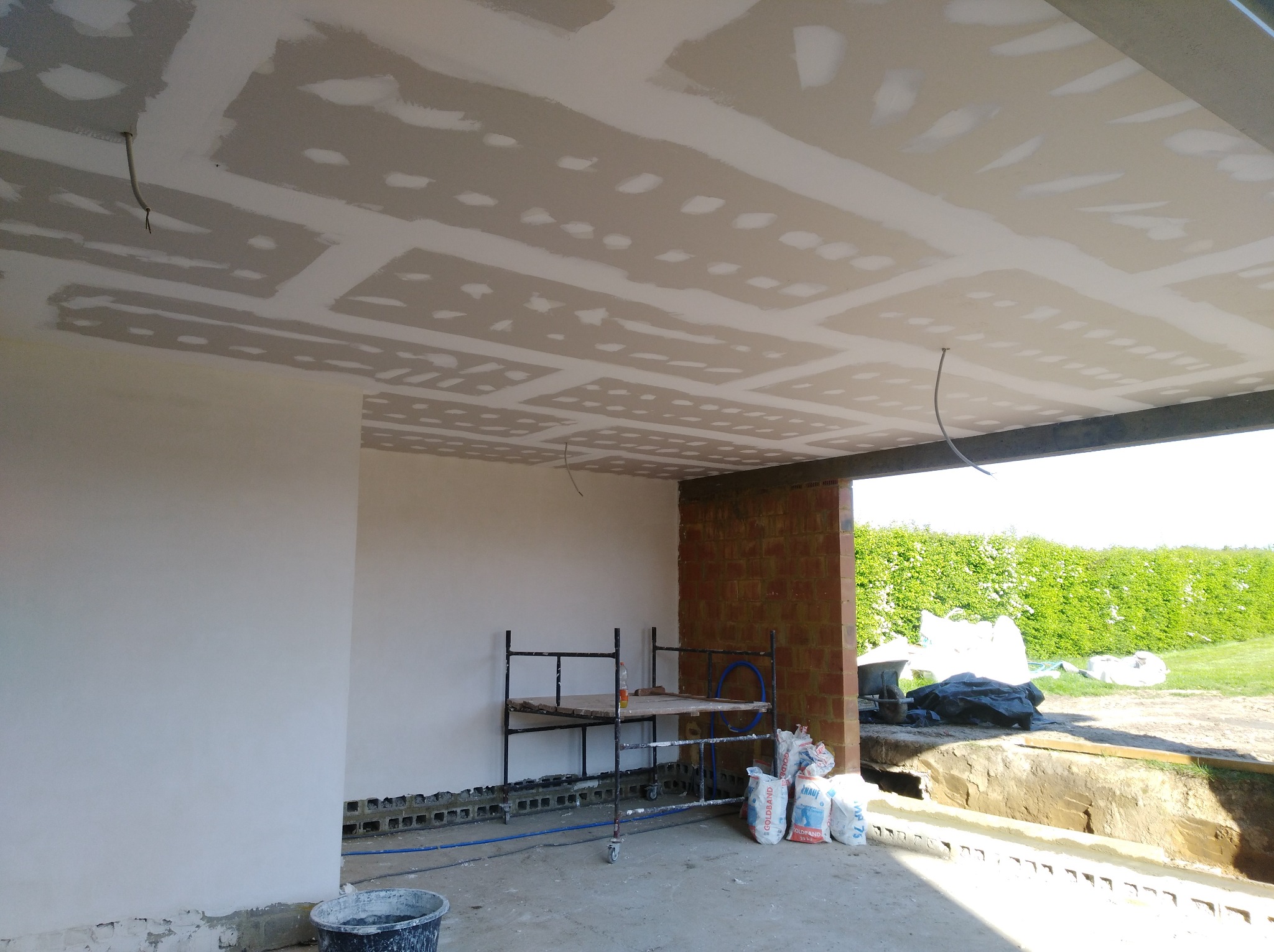 Wnętrze w trakcie prac wykończeniowych: sufit z płyt karton-gips, częściowo wyszpachlowane ściany, rusztowanie i worki z zaprawą. Widok na zewnątrz przez otwartą przestrzeń.