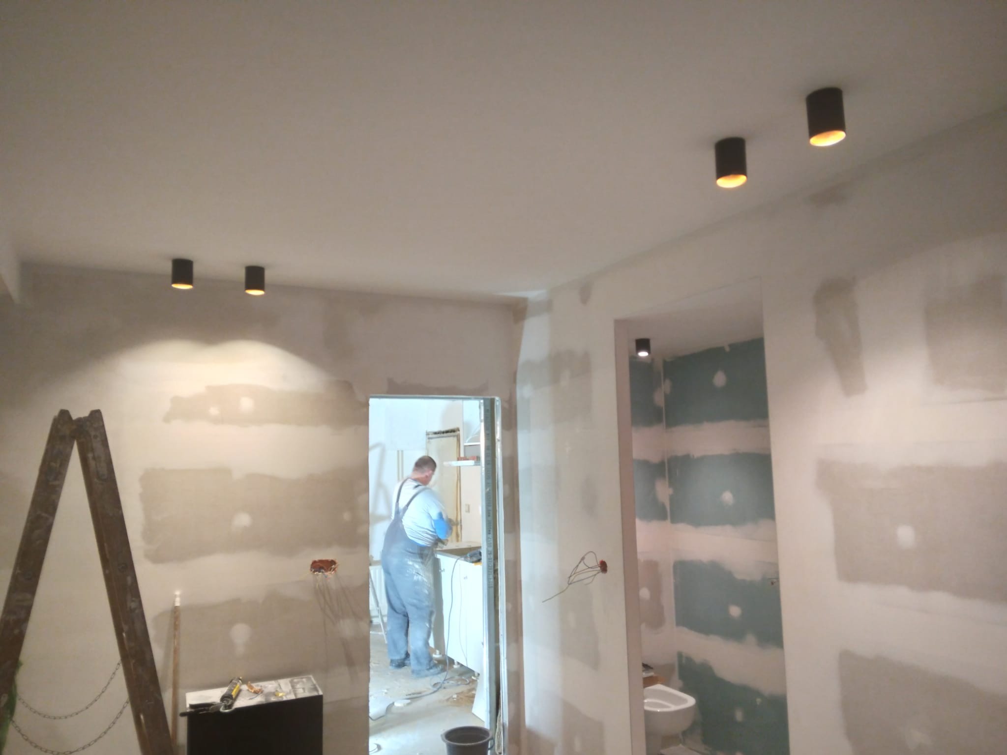 Wnętrze w trakcie prac wykończeniowych: ściany przygotowane pod malowanie, widoczne punkty oświetleniowe, drabina i pracownik w tle. Wnętrze w stanie surowym.