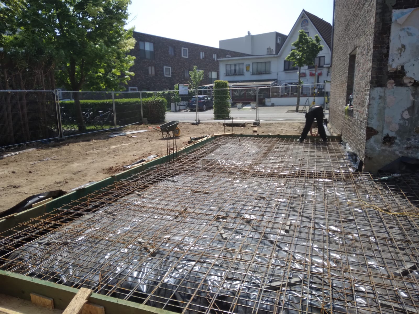 Zbrojenie fundamentów pod budowę domu jednorodzinnego. Siatka zbrojeniowa na folii, drewniane szalunki, widoczny fragment budynku w tle. Prace przygotowawcze.