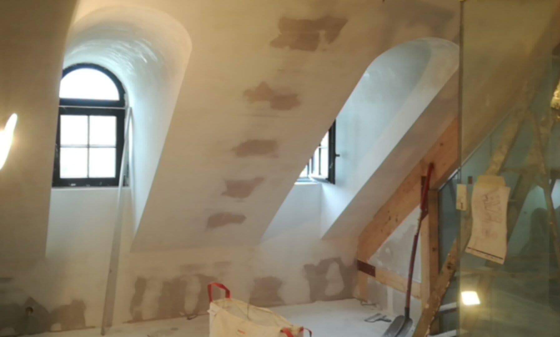 Skośne poddasze ze świeżo nałożonymi gładziami, widoczne łaty na suficie i ścianach. Okna łukowe w czarnych ramach. Przy ścianie narzędzia i torba budowlana.