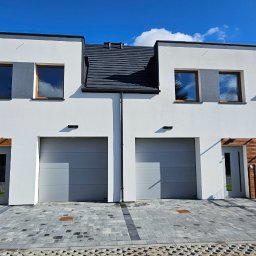 Monibud - Nowoczesny bliźniak z białymi ścianami, szarą dachówką i drewnianymi akcentami elewacji. Kostka brukowa na podjeździe, błękitne niebo w odbiciu okien.