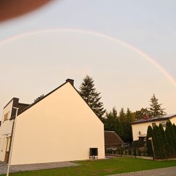 Monibud - Biała elewacja domu jednorodzinnego pod podwójną tęczą, widok z poziomu chodnika. Na pierwszym planie zaparkowany bus z reklamą firmy.