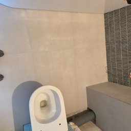 Monibud - Montaż toalety w łazience z szarą mozaiką i beżowymi płytkami. Widoczne rury i płyta gipsowa, sugerujące etap prac wykończeniowych.