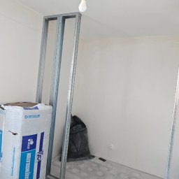 Monibud - Wnętrze w trakcie prac: metalowe profile konstrukcyjne, izolacja Knauf Insulation, wisząca żarówka. Widoczny worek na śmieci i surowe ściany. Ujęcie z dołu, podkreślające wysokość pomieszczenia.