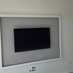 Monibud Monika Witkowska - Ściana z wnęką na telewizor, obramowaną białą ramą i wykończoną szarą gładzią. Widoczne gniazdka elektryczne i fragment okna po lewej stronie.