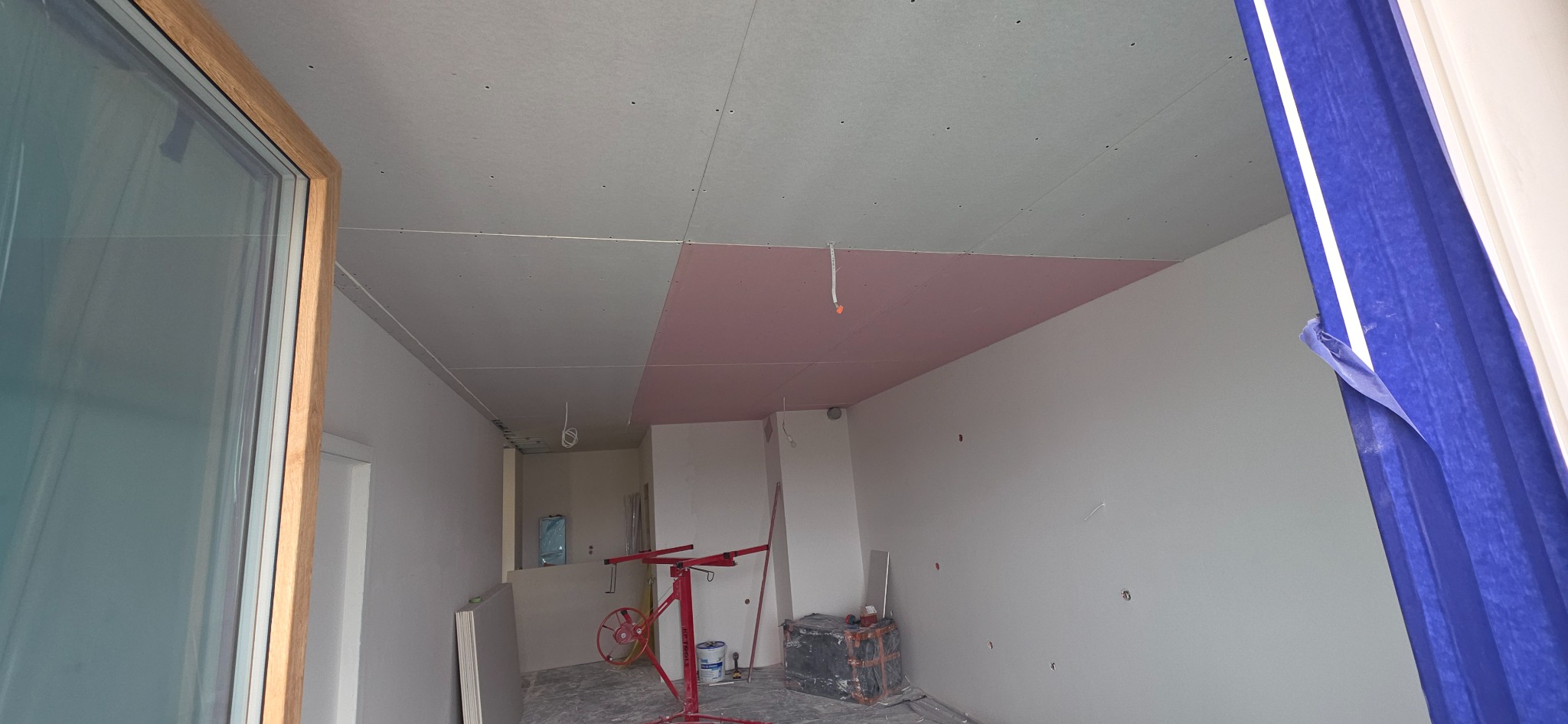 Wnętrze w trakcie montażu płyt gipsowo-kartonowych na suficie, widoczny podnośnik, częściowo zakryte okno z drewnianą ramą i niebieska folia ochronna.