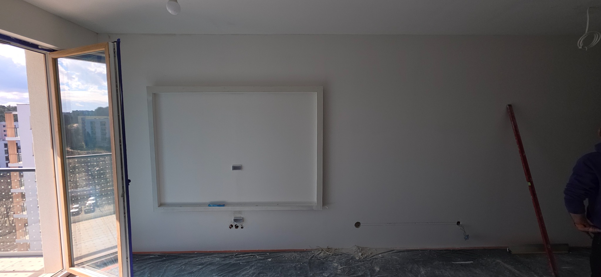 Biała ściana z przygotowaną ramą pod TV i instalacją elektryczną, widok z otwartym oknem balkonowym. Przygotowanie do wykończenia wnętrza.