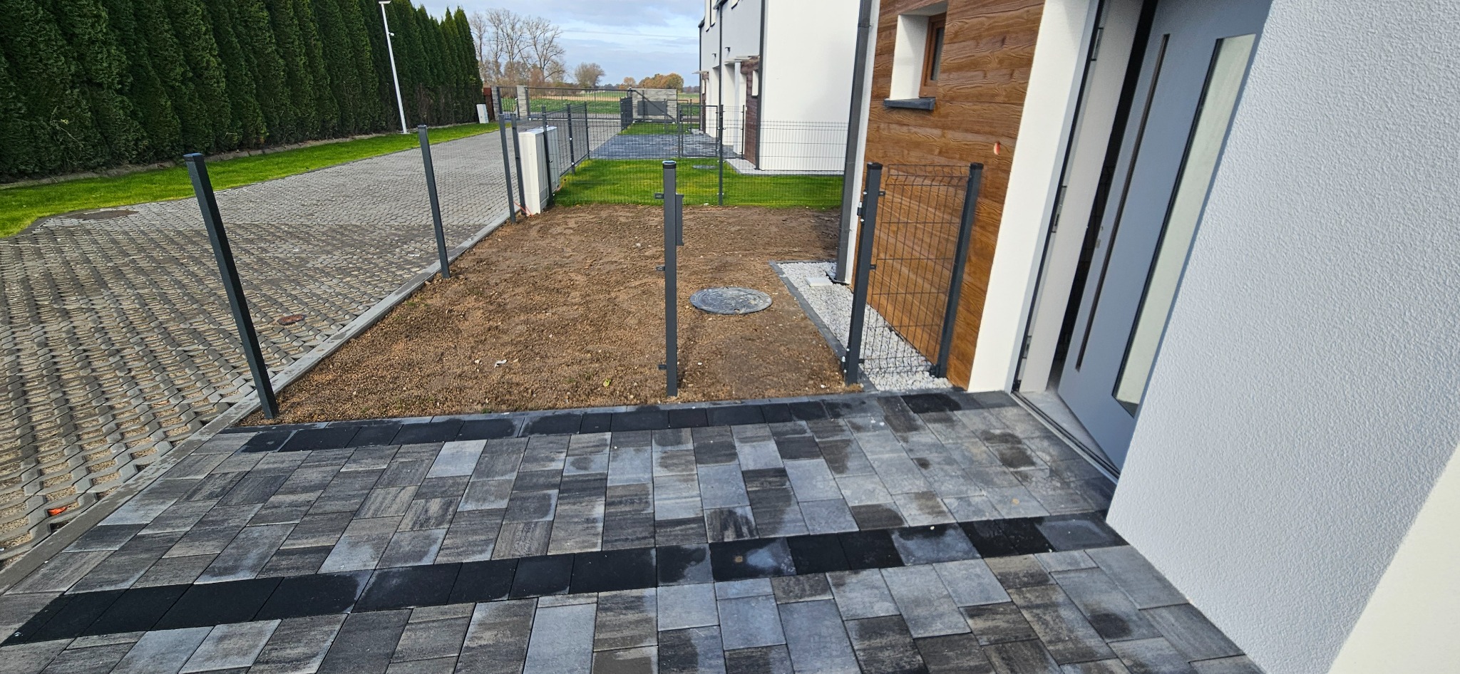 Ułożona kostka brukowa w odcieniach szarości prowadząca do wejścia domu z drewnianą elewacją. Widoczny fragment ogrodzenia i przygotowany teren pod ogród.