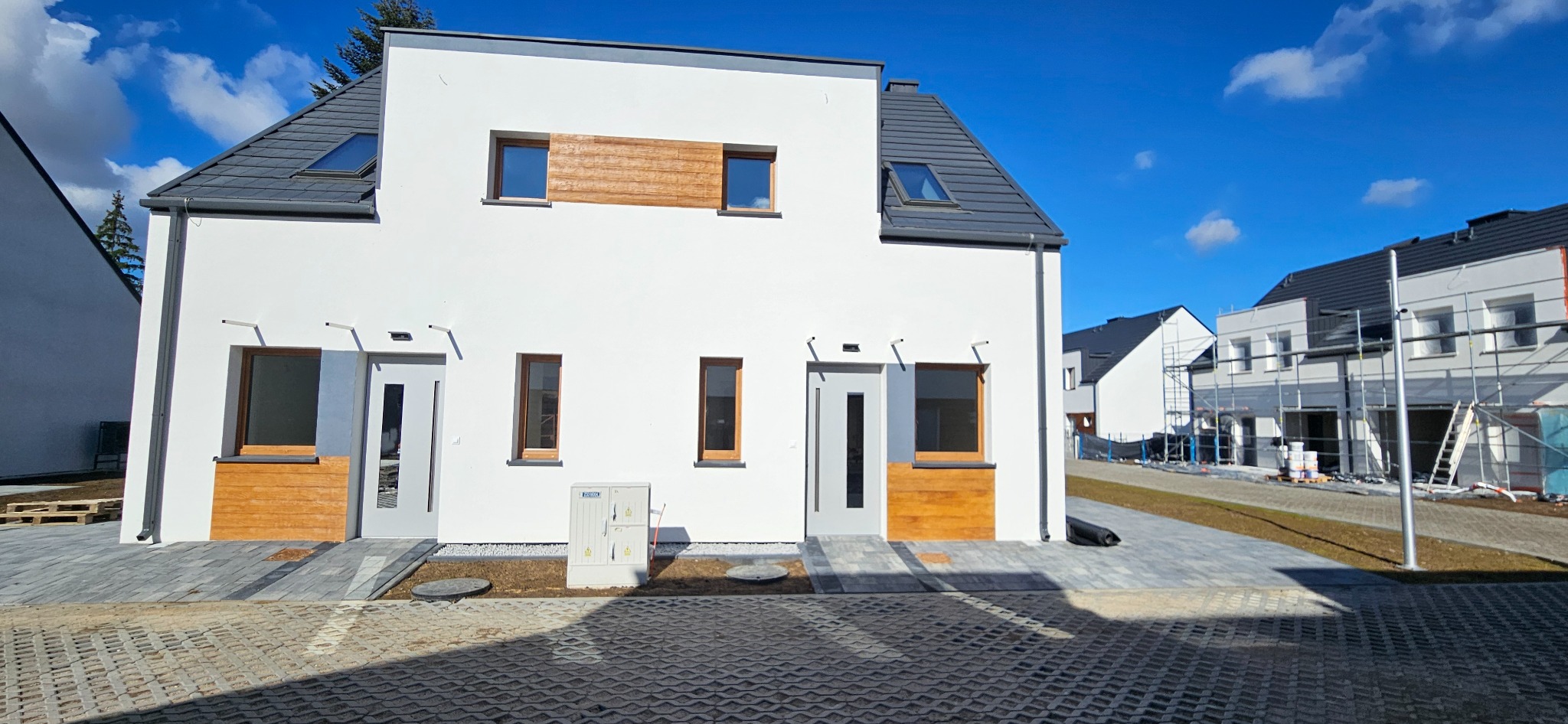 Nowoczesny bliźniak z białymi elewacjami, szarą stolarką i drewnianymi akcentami. Kostka brukowa na podjeździe, błękitne niebo w tle. Budynek po wykończeniu.