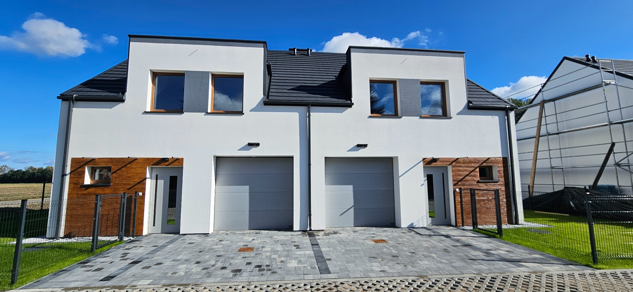 Nowoczesny bliźniak z białymi ścianami, szarą dachówką i drewnianymi akcentami elewacji. Kostka brukowa na podjeździe, błękitne niebo w odbiciu okien.