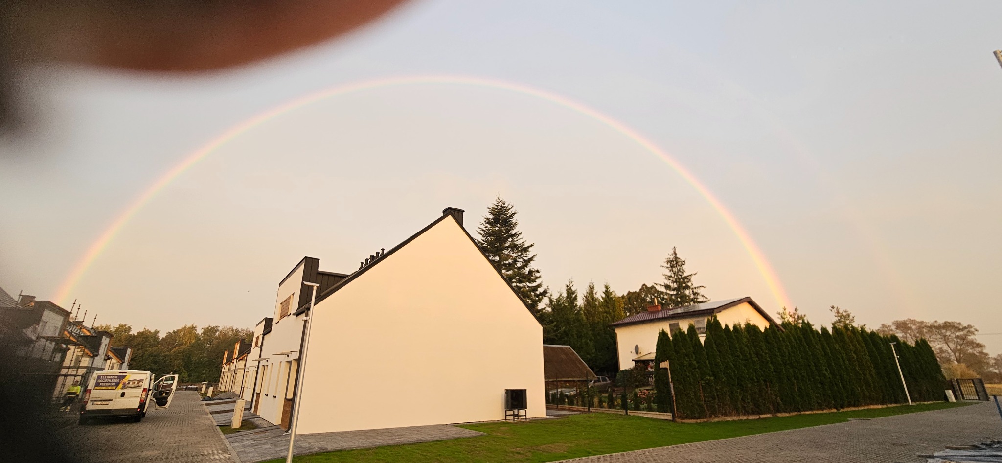 Biała elewacja domu jednorodzinnego pod podwójną tęczą, widok z poziomu chodnika. Na pierwszym planie zaparkowany bus z reklamą firmy.