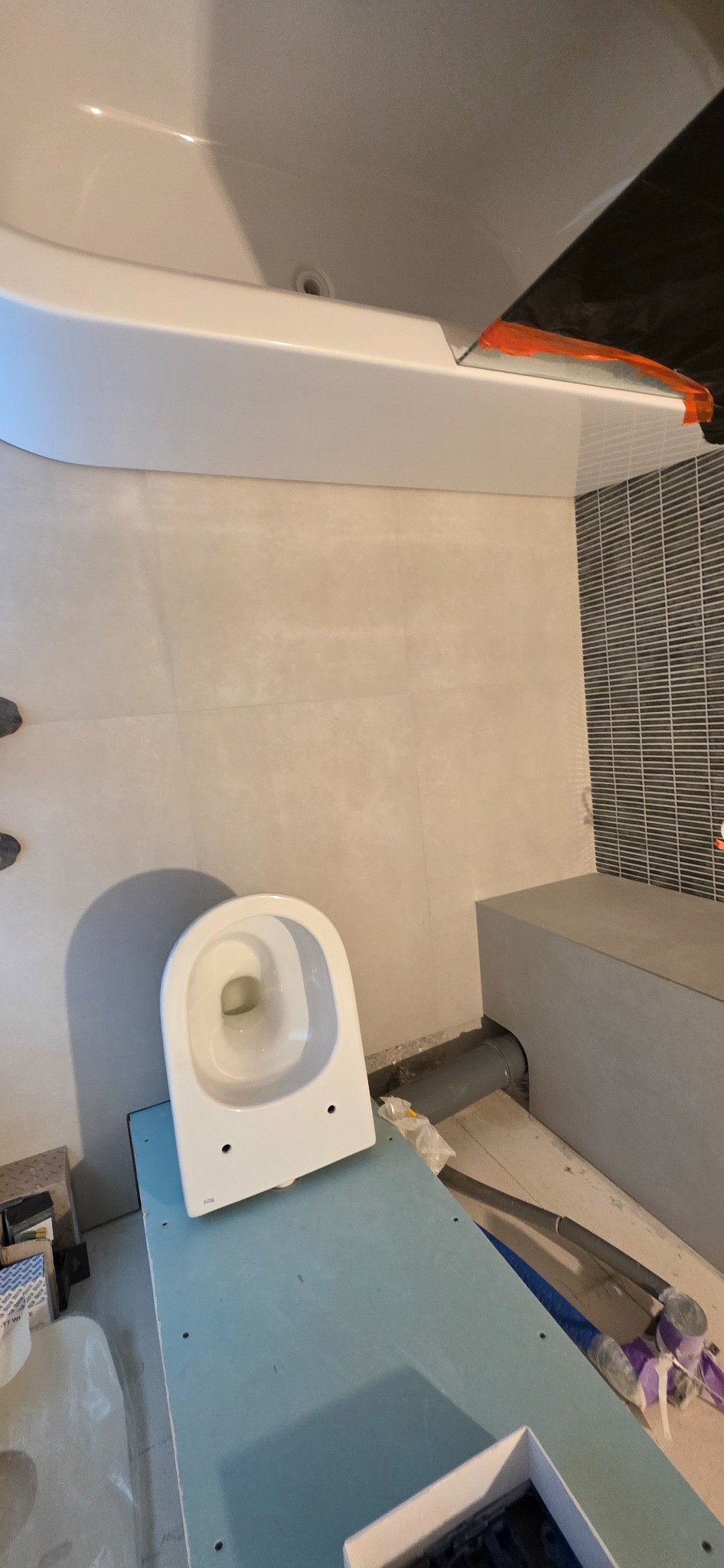 Montaż toalety w łazience z szarą mozaiką i beżowymi płytkami. Widoczne rury i płyta gipsowa, sugerujące etap prac wykończeniowych.