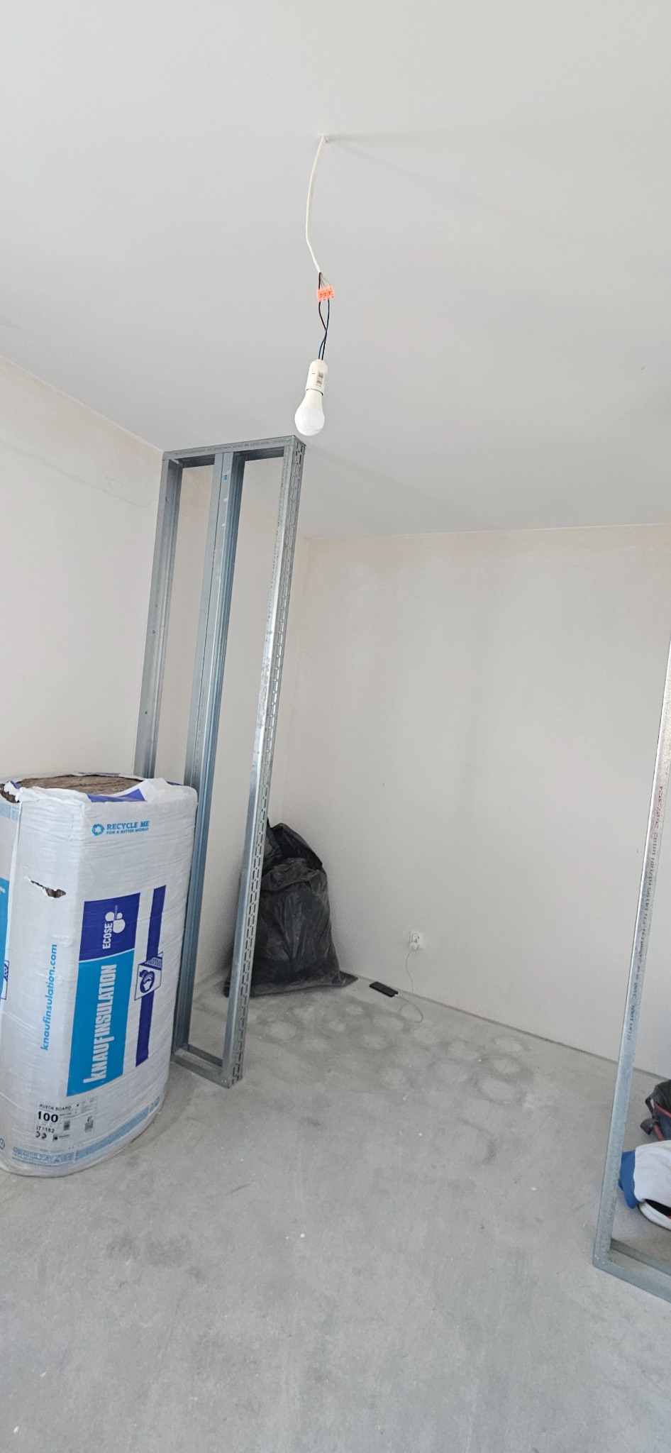 Wnętrze w trakcie prac: metalowe profile konstrukcyjne, izolacja Knauf Insulation, wisząca żarówka. Widoczny worek na śmieci i surowe ściany. Ujęcie z dołu, podkreślające wysokość pomieszczenia.