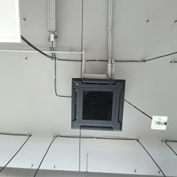 El-Klimbud Szymon Jerzycki - Sufitowa klimatyzacja kasetonowa w biurze, widok z dołu. Widoczne przewody i rury instalacyjne. Szara obudowa klimatyzatora z kratką wentylacyjną.