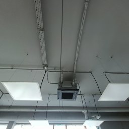 El-Klimbud Szymon Jerzycki - Sufit biurowy z zamontowaną kasetonową klimatyzacją, widoczne kable, rury i oświetlenie LED. Ujęcie z dołu, podkreślające instalacje.