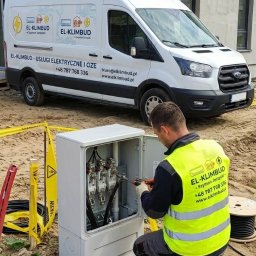 El-Klimbud Szymon Jerzycki - Pracownik w kamizelce odblaskowej łączy kable w skrzynce elektrycznej na tle budynku w budowie i firmowego busa z logo El-Klimbud.