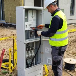 El-Klimbud Szymon Jerzycki - Elektryk w kasku i kamizelce łączy kable w otwartej skrzynce elektrycznej na zewnątrz budynku w trakcie budowy. Widoczne narzędzia i zwój kabla.