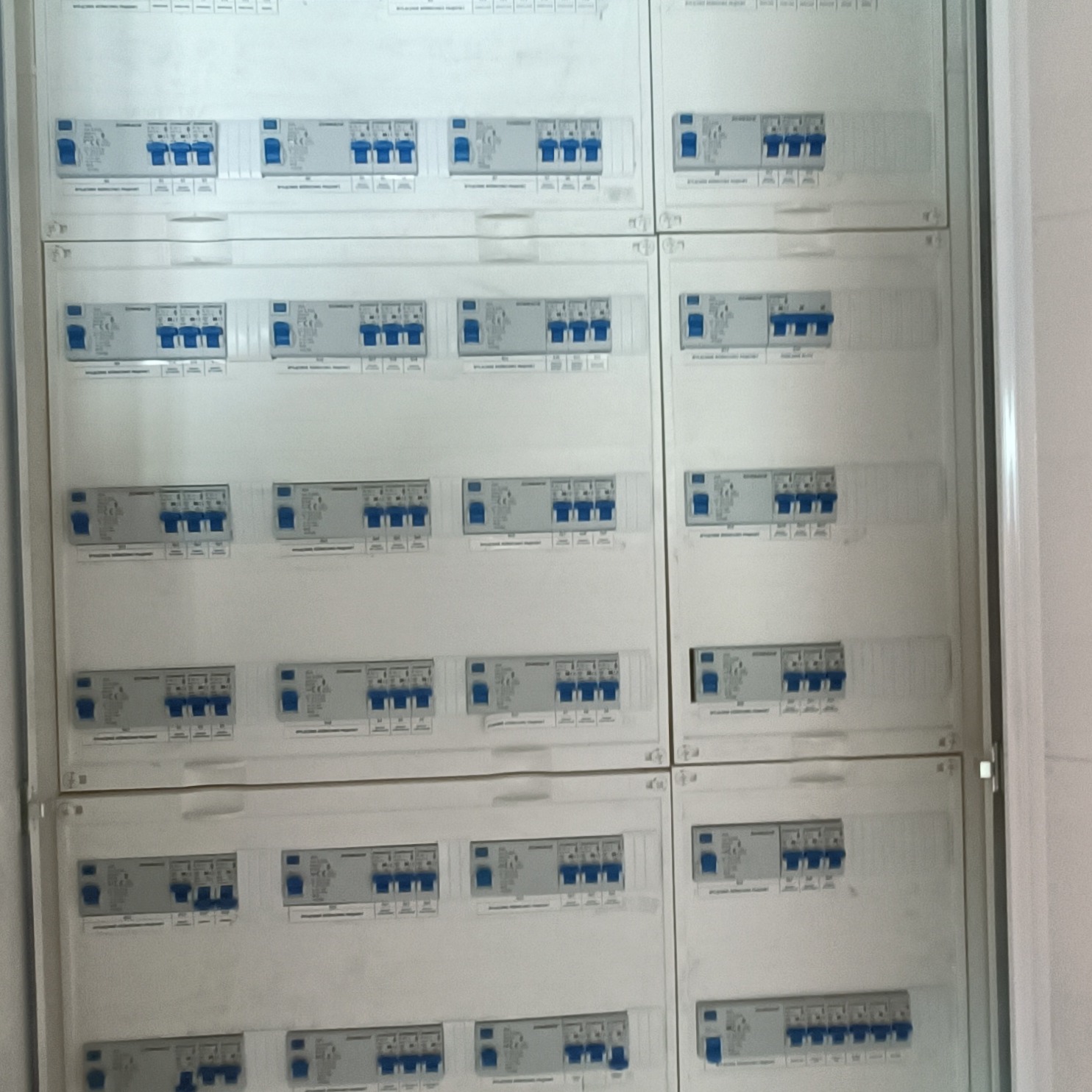 Rozdzielnia elektryczna z wieloma bezpiecznikami, widok z bliska. Biała obudowa, niebieskie przełączniki. Układ symetryczny, równomierne oświetlenie.