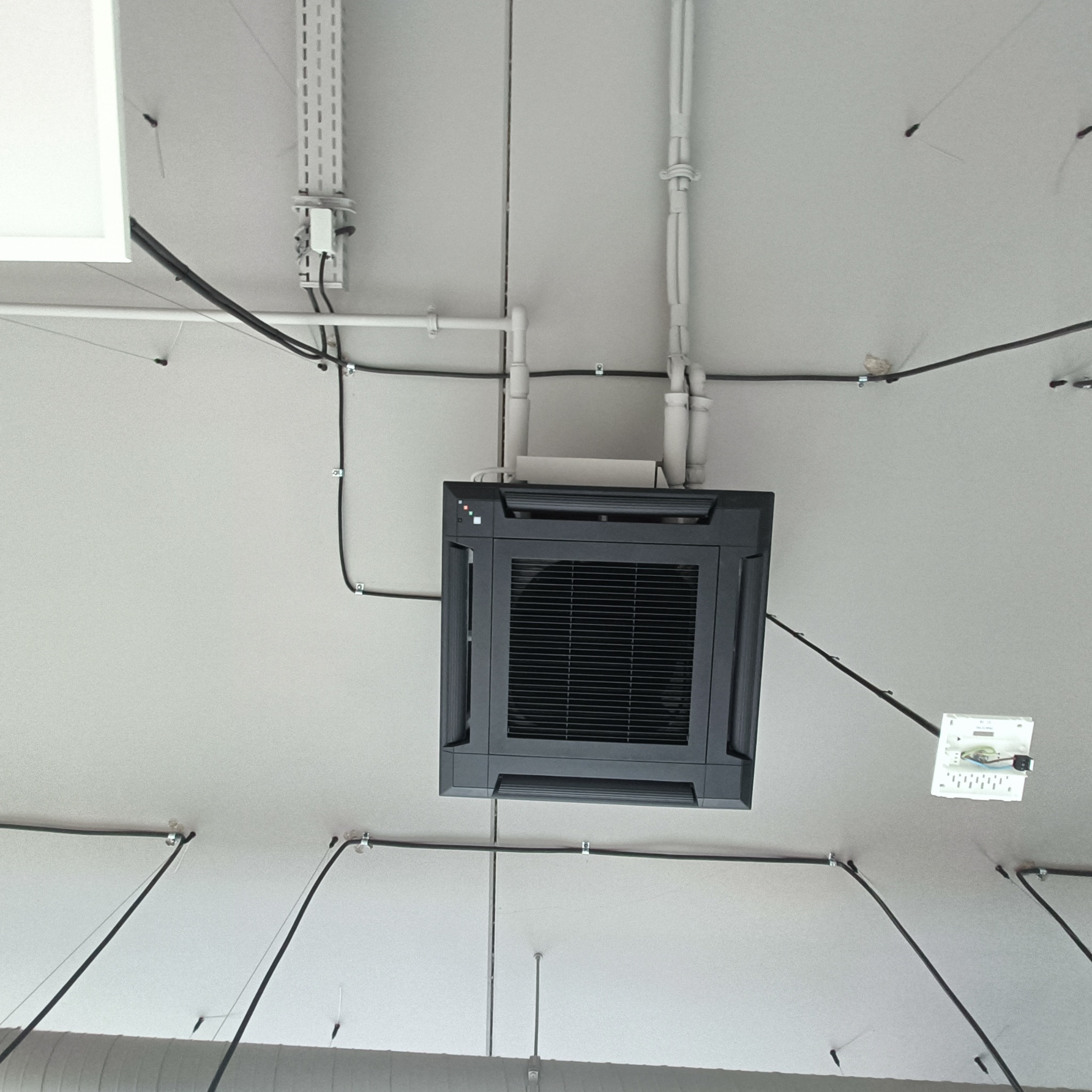Sufitowa klimatyzacja kasetonowa w biurze, widok z dołu. Widoczne przewody i rury instalacyjne. Szara obudowa klimatyzatora z kratką wentylacyjną.