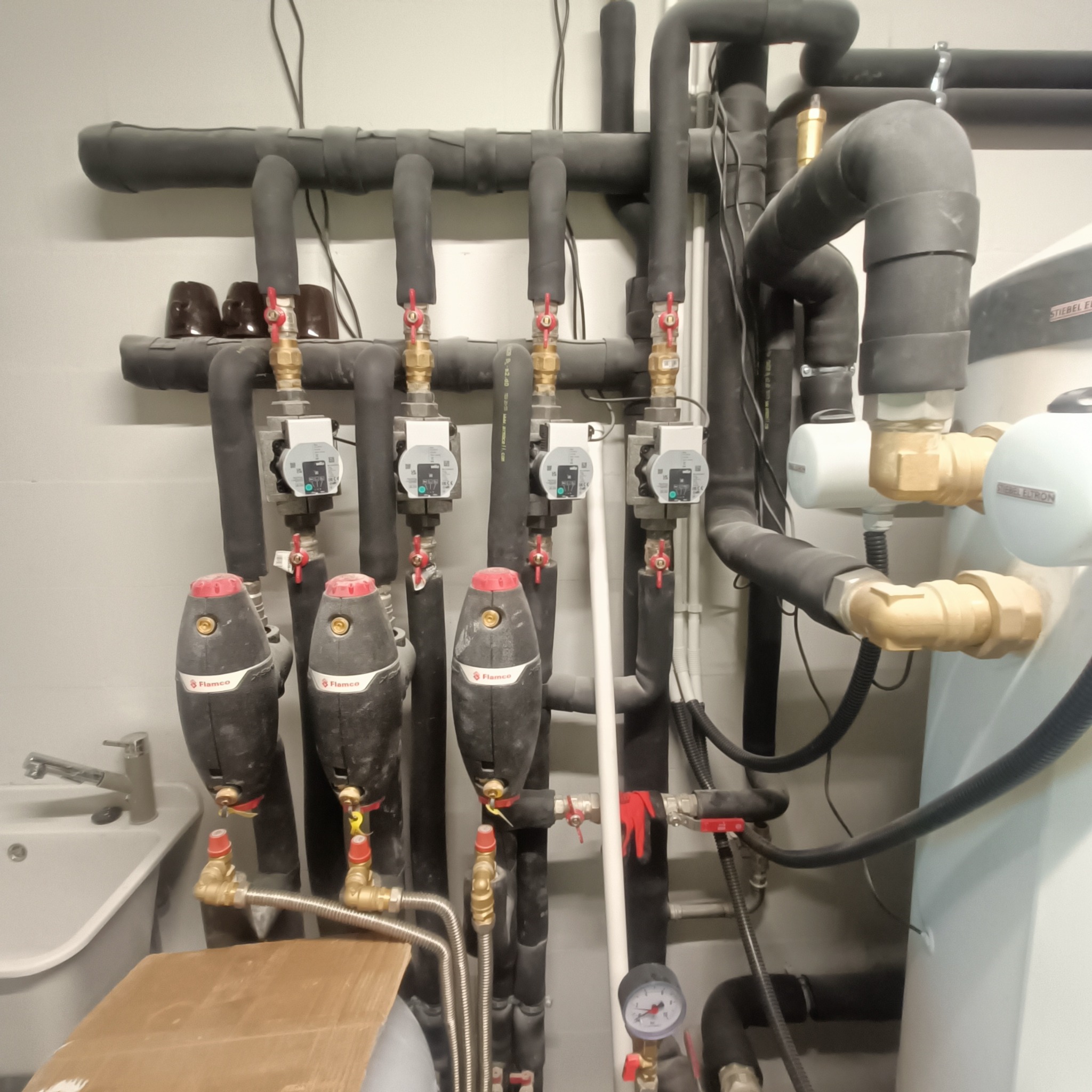 Złożona instalacja grzewcza z czarnymi rurami, zaworami z czerwonymi pokrętłami i białym zbiornikiem Stiebel Eltron. Widoczny manometr i elementy hydrauliczne.