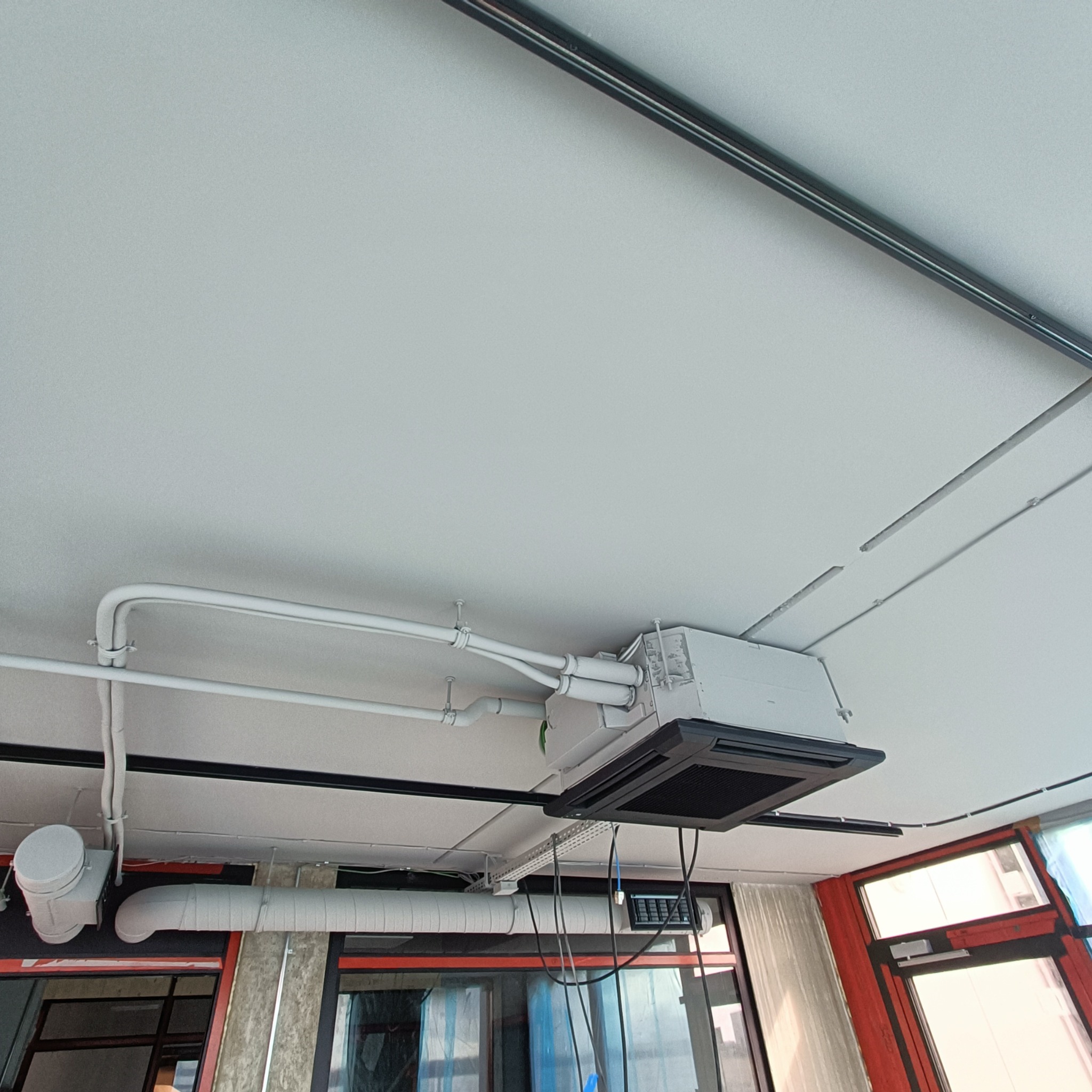 Zamontowana kasetonowa klimatyzacja sufitowa w biurze, widoczne rury i kable. Białe elementy instalacji na tle jasnego sufitu. Ujęcie z dołu, widoczne okna.