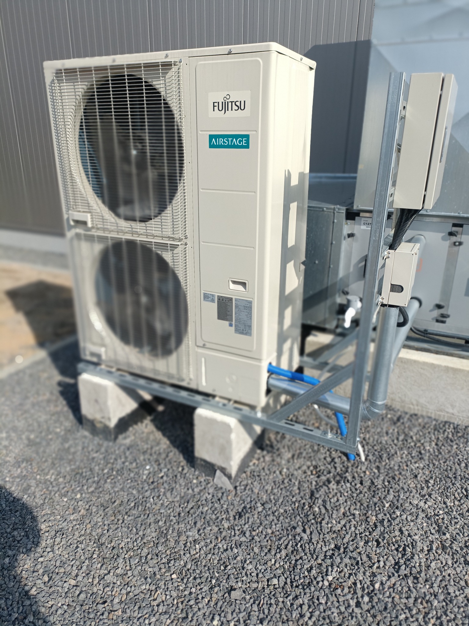 Zewnętrzna jednostka klimatyzacji Fujitsu Airstage na betonowych podstawach, z widocznymi niebieskimi rurami i metalową konstrukcją wsporczą na tle szarej ściany.
